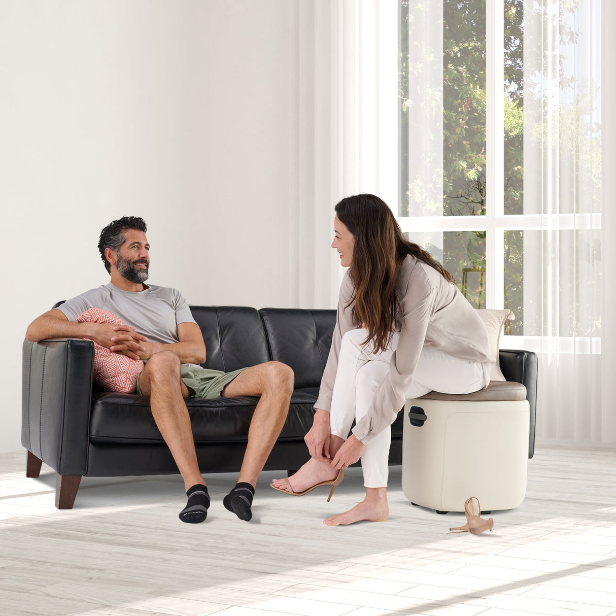 Reveal Ottoman Massager shown in bright living room with person relaxing on couch while partner demonstrates foot massage feature